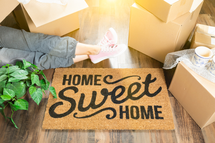 Woman Wearing Sweats Relaxing Near Home Sweet Home Welcome Mat, Moving Boxes and Plant. Woman Wearing Sweats Relaxing Near Home Sweet Home Welcome Mat, Moving Boxes and Plant.
